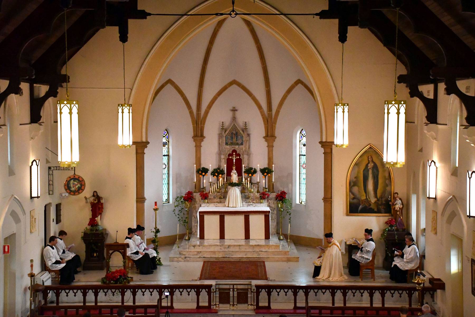 The sanctuary of a church during a Traditional Latin Mass: the altar under a pointed arch, with the priest and servers seated at either side.